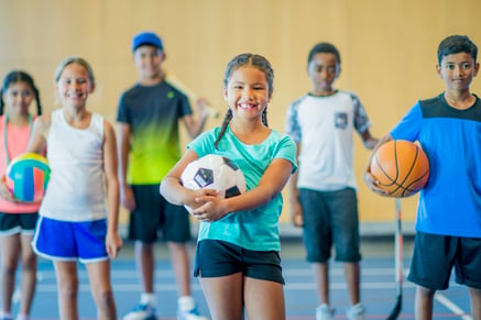 A line of six children with one at the forefront. Each are holding a sport ball such as volleyball, soccer, and basketball. 