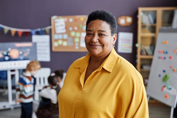 A woman teacher of a child care centre looks at the camera while children play in the background. 