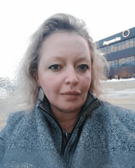 A ‘selfie’ of a person in a grey sweater, taken from the shoulders up. We can see grass in the background, as well as an orange and black building.