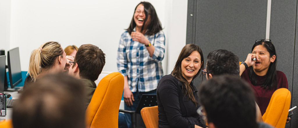 A Payworks employee stands in the middle of a group of seated coworkers during a seminar while they all share a laugh. 