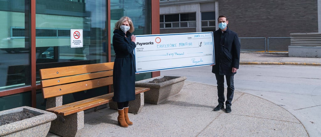 Patti Smith, President and CEO of CancerCare Manitoba Foundation with Payworks President & CEO JP Perron outside of the CancerCare Manitoba building in downtown Winnipeg. They’re holding a large cheque for $50,000 and are both wearing masks.  