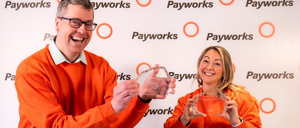 Two Payworks staff in matching orange sweatshirts holding up two awards accompanied by text that reads, “We’re Canada’s Top Small & Medium Employer for 2024!”.  