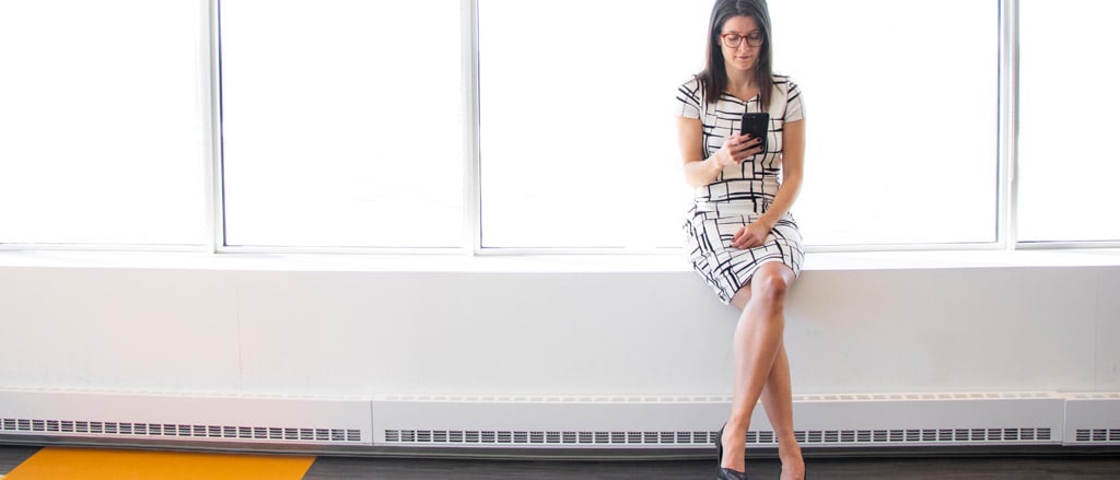 A woman sitting on a ledge inside of an office building with all windows in the background. She is looking down at her cell phone.  