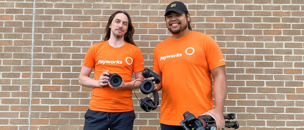 Deux employés de Payworks vêtus de t-shirts orange assortis et qui manient de l’équipement de vidéaste, accompagnés du texte « Pay it Forward » (Donner au suivant). 