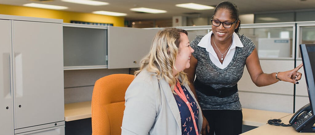 Une femme pointant un écran d’ordinateur dans le cubicule d’une autre personne, accompagnée du texte suivant : « Semaine du service à la clientèle : retour sur l’histoire de notre service ». 