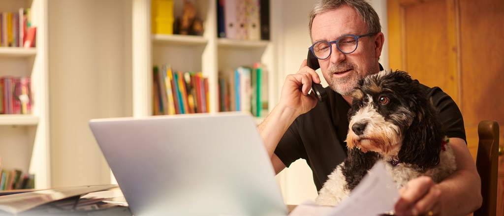 An older man with glasses sitting at a home office desk with a laptop open in front of him and a bookshelf behind him. He’s on the phone and is looking down at some papers on his desk. There’s also a black and white dog sitting on his lap looking off into the distance.  