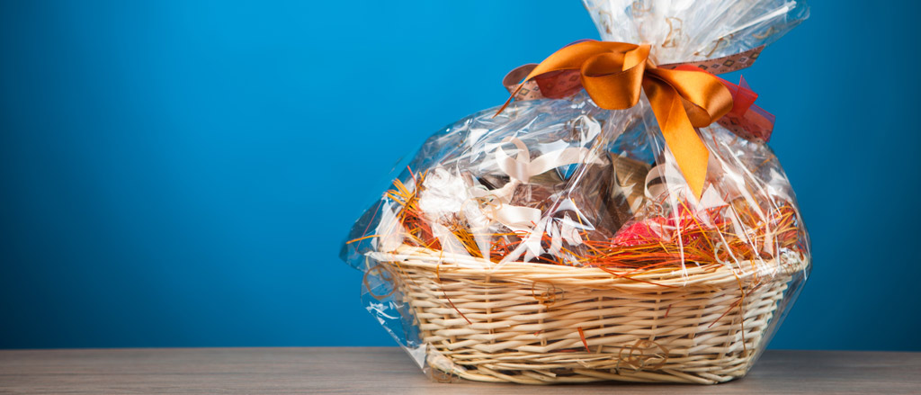 A wicker gift basket with lots of goodies inside it, wrapped in cellophane wrapping and an orange ribbon tied into a bow. The gift basket is on a wooden table and there is a sky blue background behind it.  