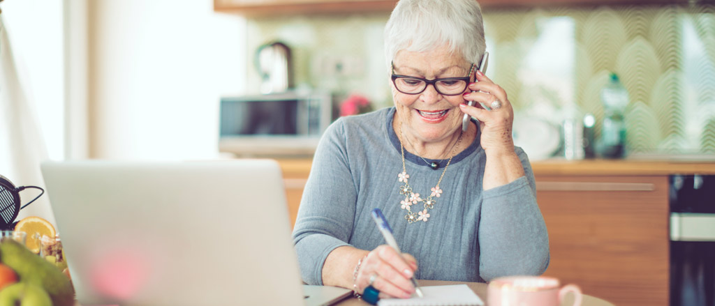 An elderly lady in a blue long sleeve shirt and dark rimmed sunglasses, listening to a phone call on her cell phone and taking notes on a scribbler in front of her. She's at her home office with a kitchen in the background and an open silver laptop on the left-hand side. 