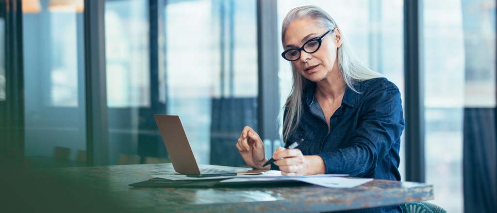 Une femme travaille sur un ordinateur portable. Elle regarde un document papier. Il est écrit : « Liste exhaustive des jours fériés en 2022 ». 