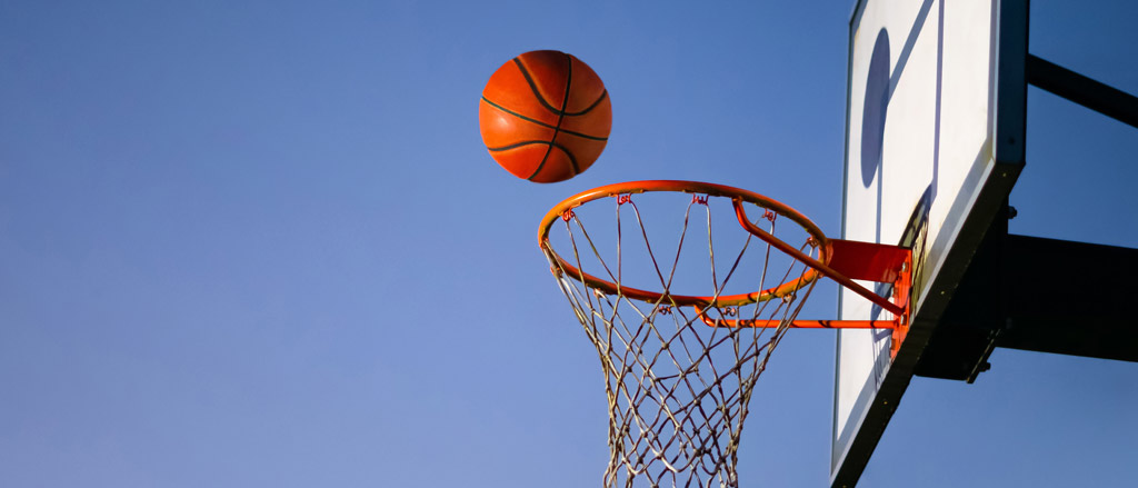 A basketball net off to the left-hand side with a clean blue sky in the background. An orange basketball is in midair, right above the hoop about to swoosh in.  
