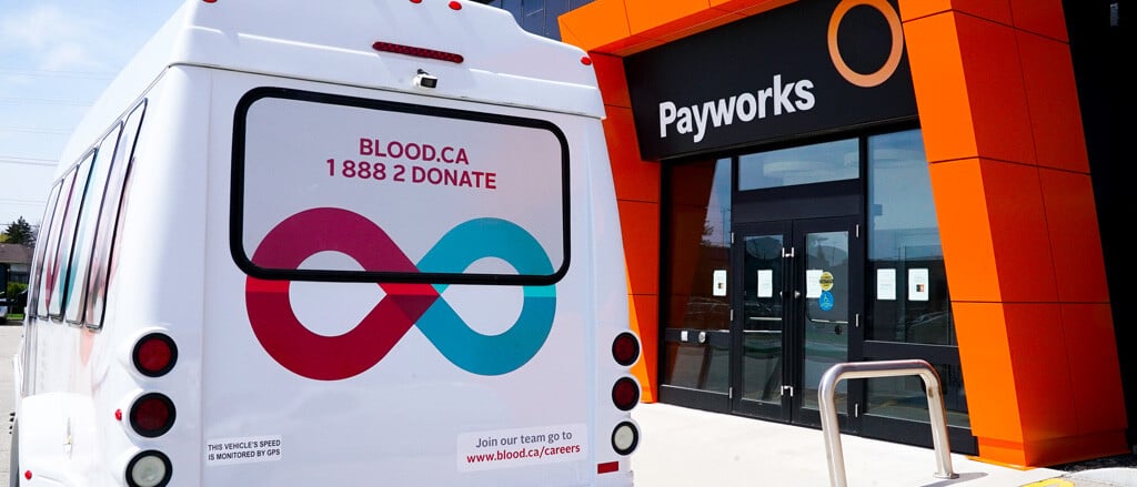 The front of the Payworks HQ office with a Canadian Blood Services transportation van parked in front. Text reads, “Pay it Forward”.  