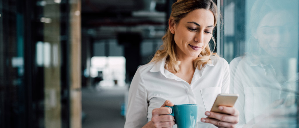 A woman leaning against a glass window of an office holding a teal coffee cup and looking down at her cell phone.  