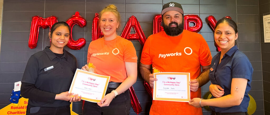 Deux employés de Payworks vêtus de t-shirts orange qui reçoivent des certificats de bénévolat, accompagnés du texte « Pay it Forward » (Donner au suivant). 