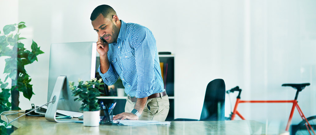 A man standing up from his chair at the office. He has a desktop computer in front of him. He has his mobile phone up to his ear and one hand on the desk ready to type.  