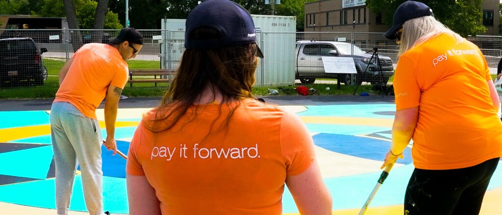 Deux employés de Payworks qui aident à peindre un terrain de basketball, accompagnés du texte « Pay it Forward » (Donner au suivant).