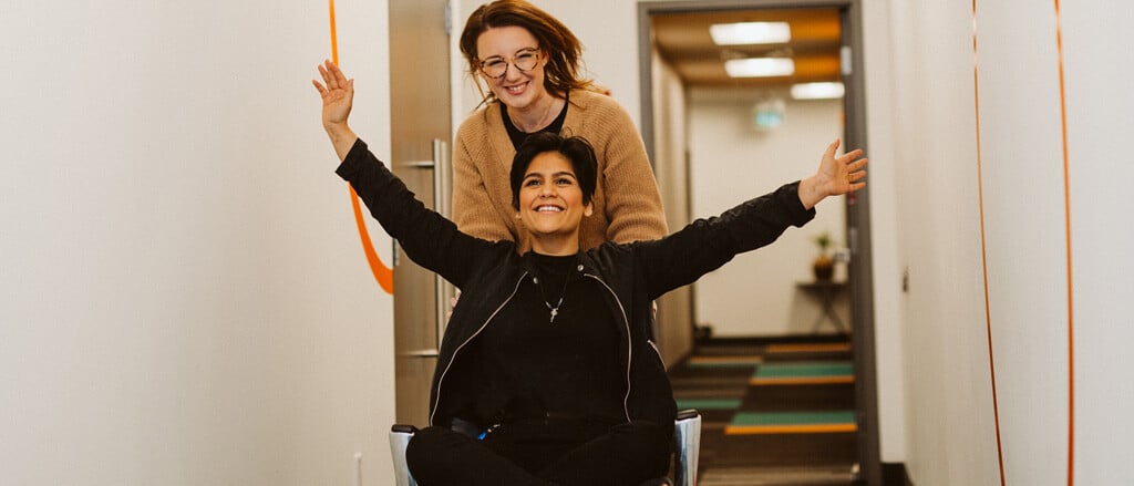 An individual pushing another on a chair down a hallway accompanied by text that reads, “our top reasons to thank your own payroll start this week!”. 