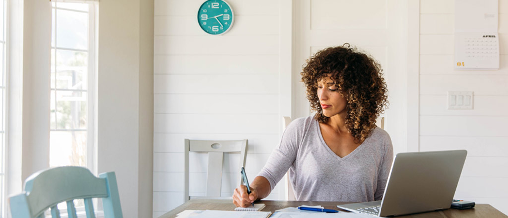 A lady sitting at her kitchen table working with a laptop and a bunch of papers in front of her. 