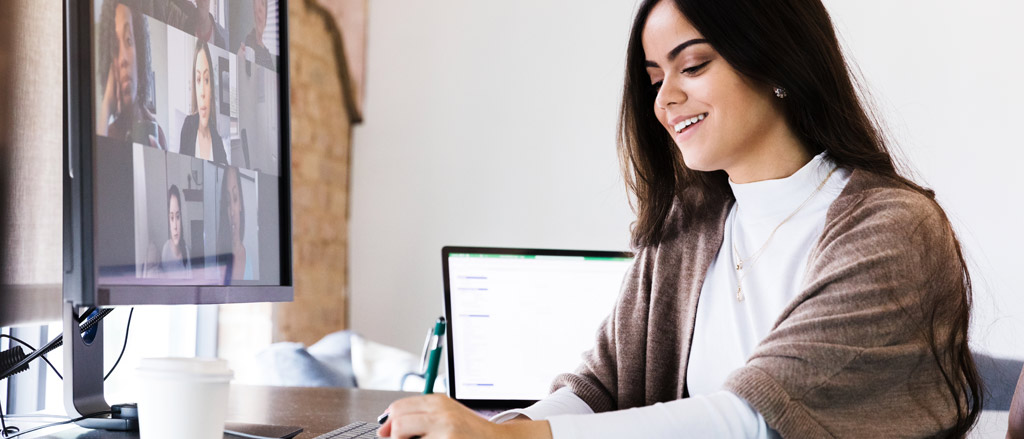 A young professional woman sitting at a home office desk with an open laptop to her right hand side and a large monitor with a video call on in front of her. She’s taking notes on a piece of paper in front of her and is looking down and smiling.  