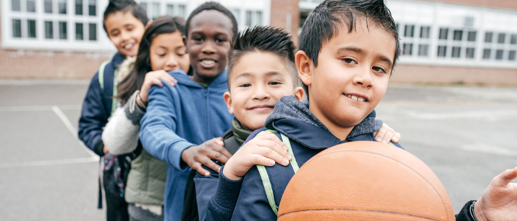Five kids standing in a line with their hands on each other’s shoulders. The child at the front of the line is holding a basketball.  