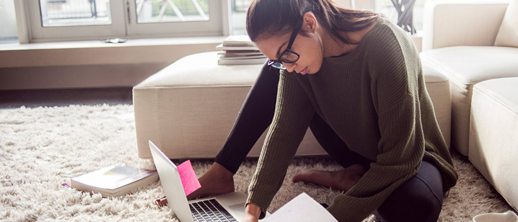 A woman wearing a sweater, tights and glasses with her hair pulled back in a pony tail, sitting on the floor of her living room with a couch in the background. She has a laptop open in front of her with pink sticky notes on the screen and a large book off to the side. She’s looking through papers off to the side of her laptop.  