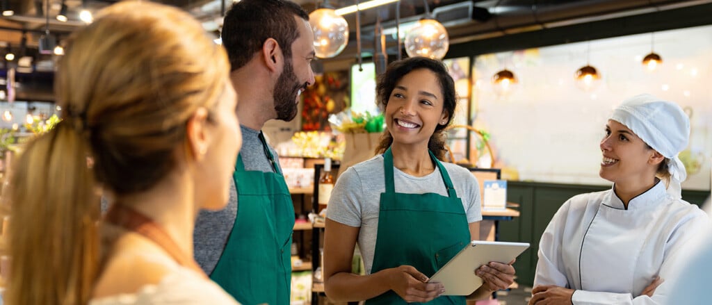 A group of four employees in a store chatting with tablets and clip boards accompanied by text that reads, “HR for small businesses: help, without the extra headcount”. 