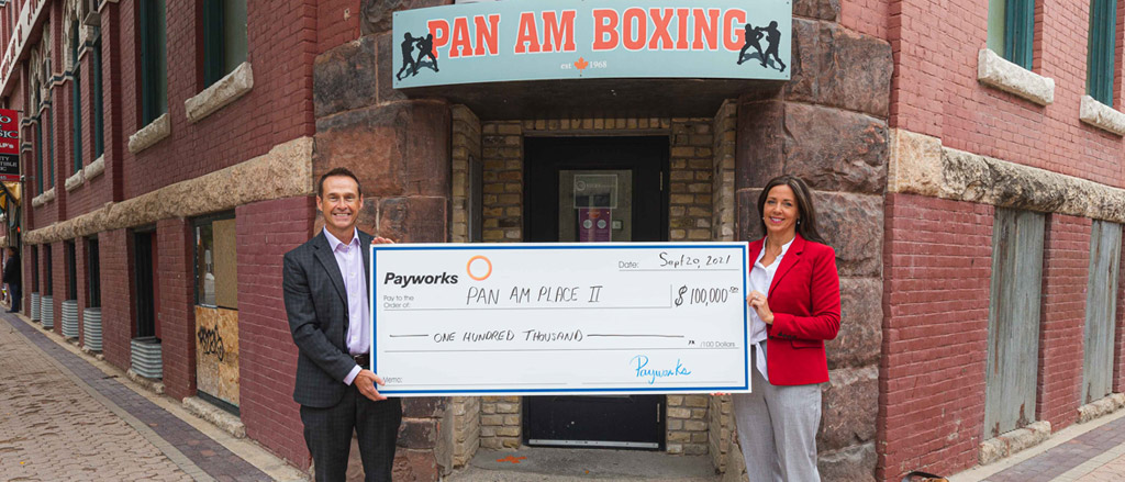 Two indivduals holding a large cheque for $100,000 under a sign that says Pan Am Boxing. Text reads, “Supporting holistic health at Pan Am Place 2”.  