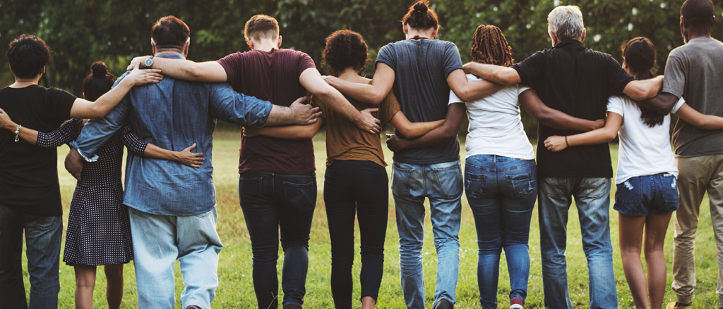 10 individuals walking through an outdoor space with grass and trees. They all have their hands around one another. Text reads, “Watch! How we celebrated two decades of paying it forward”.  