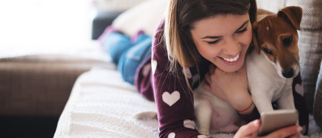 A woman sitting on her couch with her dog looking at a phone. Text reads, “the three C’s of virtual team-building”.  