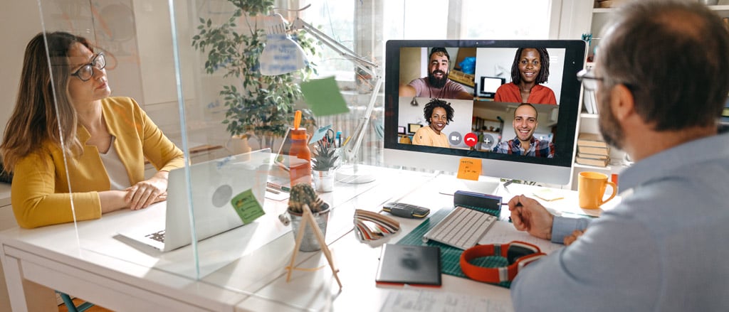 Two employees sitting at an office desk separated by a Plexiglas partition. They are looking at a video call with four people on the computer screen. Text reads “three ways to adapt your onboarding experience for remote work”.  