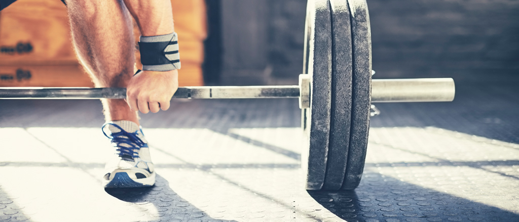 A close up photograph of on end of a barbell on a gym floor with three weights on the end. You can see an arm and a leg of a man getting ready to pick it up wearing runners and wrist supports.  