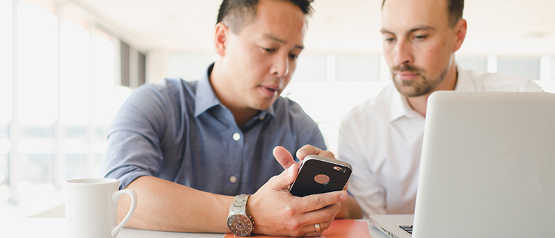 Two men sitting at a desk, looking down at a mobile phone. Text reads, “deliver employee tax forms with ease & confidence”.  